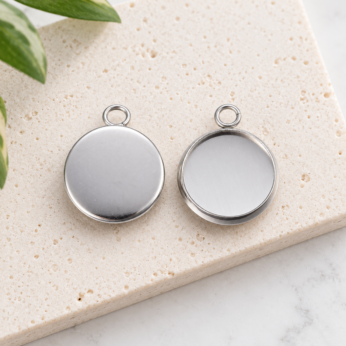Front and back view of 12mm round stainless steel pendant settings on a light stone background, showing the bezel cup and smooth reverse side.