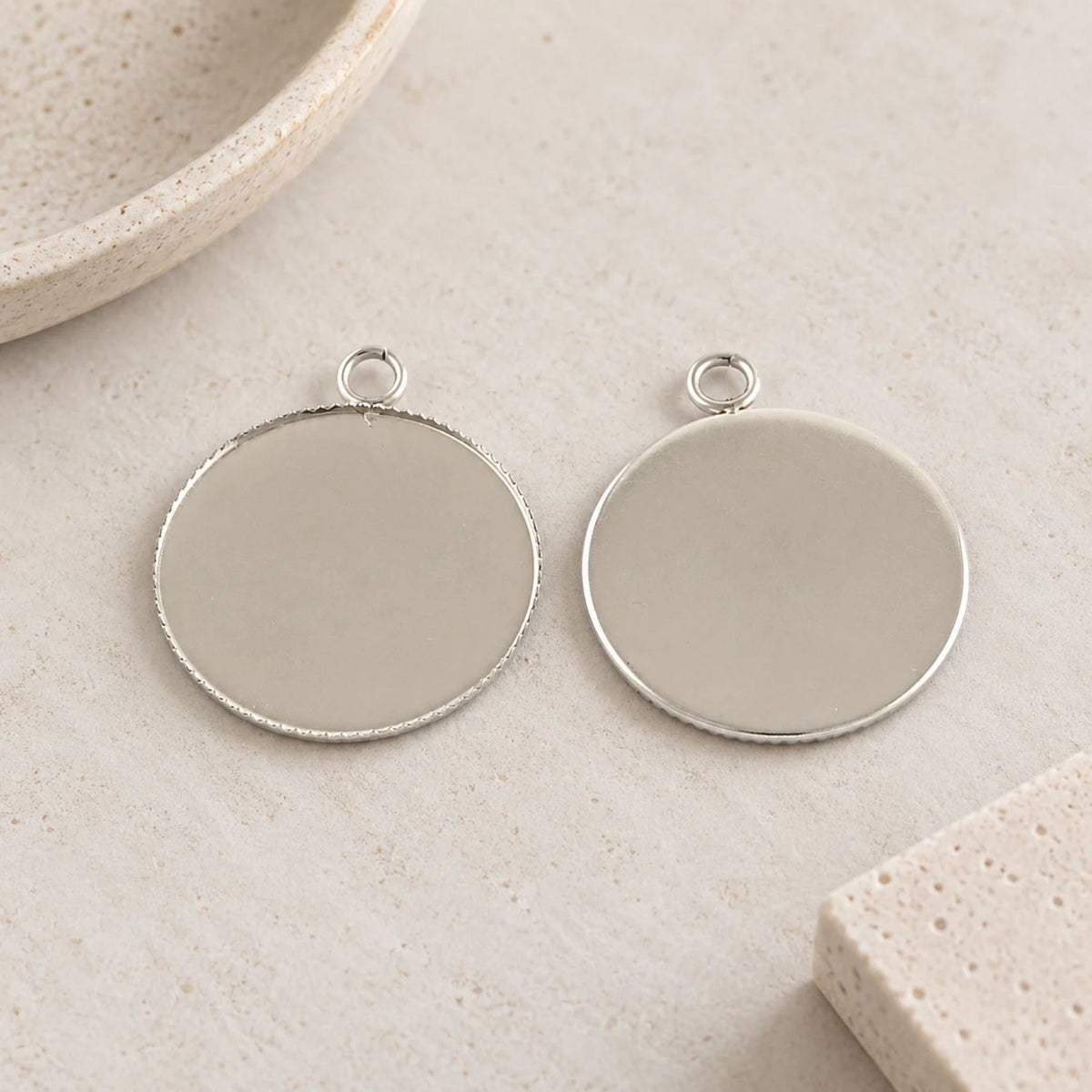 Two round 25mm stainless steel pendant settings with finely textured edges, shown from the back on a neutral beige background.