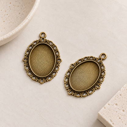 Two antique bronze oval pendant settings with decorative filigree borders, designed for 18x25mm cabochons.
