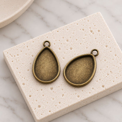 Two antique bronze teardrop pendant bezels for 18x25mm cabochons displayed on a light textured stone background.