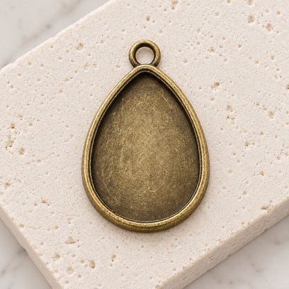 Two antique bronze teardrop pendant trays displayed on a neutral background for jewelry making.