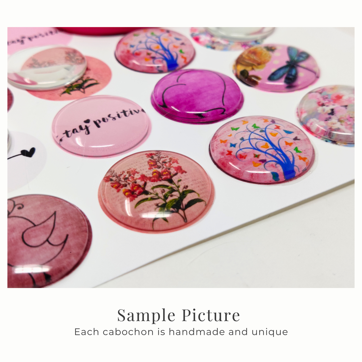 Close-up of domed round cabochon with various pink and multicolor designs, including floral, bird, and tree illustrations, displayed on a white background with text below reading 'Sample Picture Each cabochon is handmade and unique'.