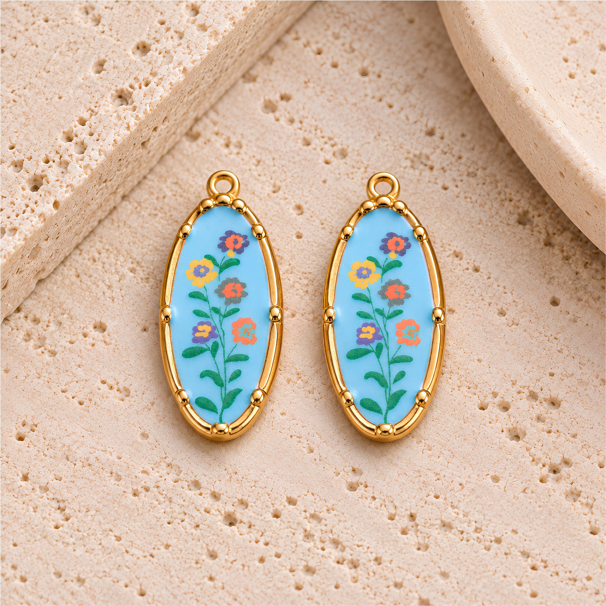 Two light blue floral oval charms with gold-plated stainless steel frames on a beige stone background.