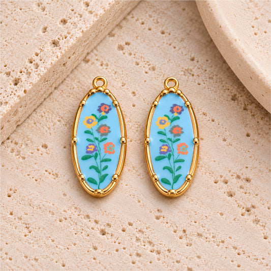 Two light blue floral oval charms with gold-plated stainless steel frames on a beige stone background.