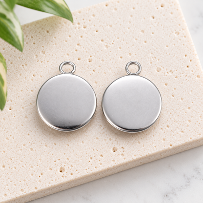 Two small round stainless steel pendant blanks showing their smooth silver backs and attachment loops on a neutral stone surface.