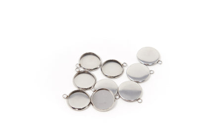 Two small round stainless steel pendant blanks showing their smooth silver backs and attachment loops on a neutral stone surface.